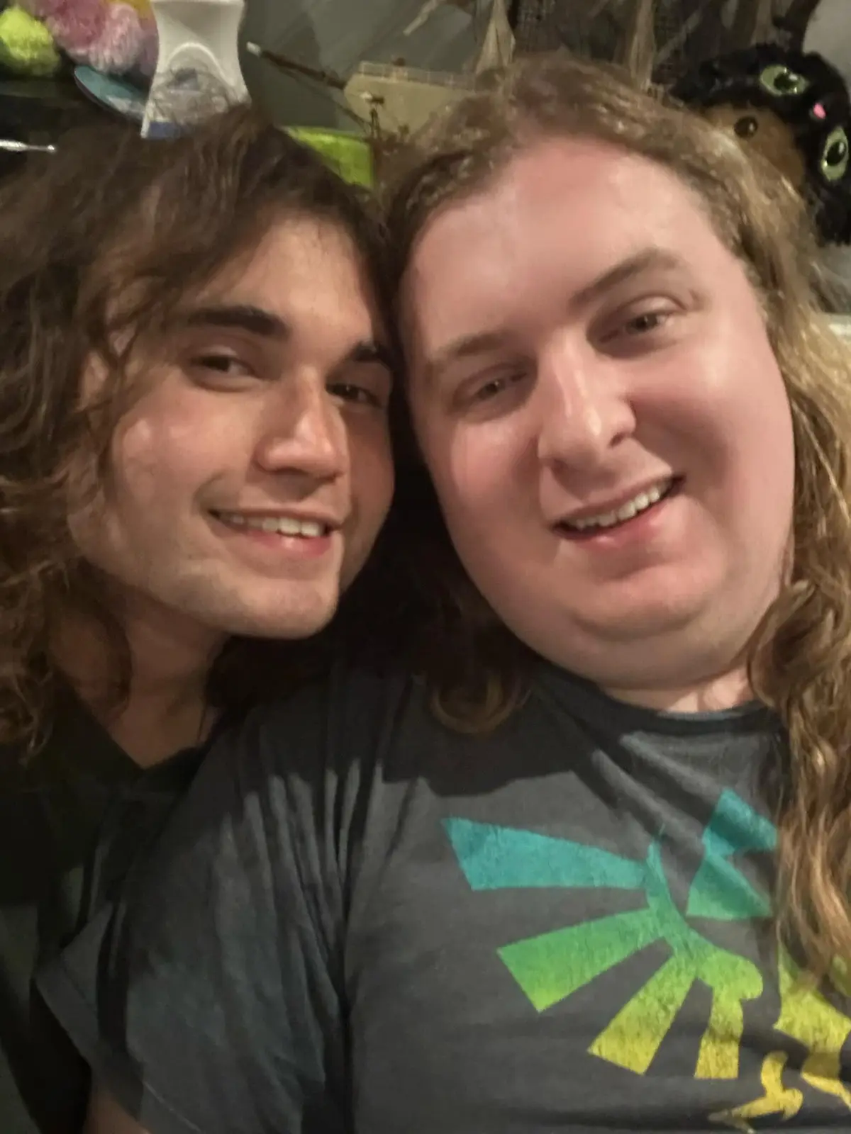 Two men with long brown hair are smiling closely together in a selfie. The man on the right wears a gray t-shirt with a…