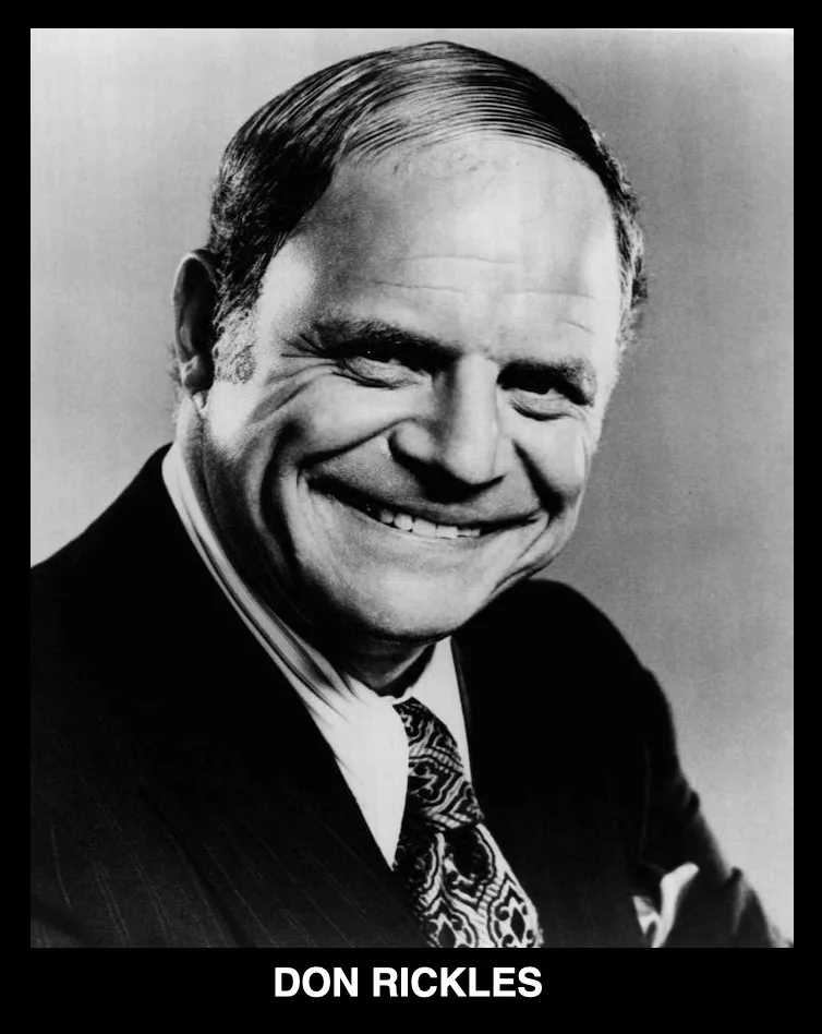 A black and white portrait of comedian Don Rickles, smiling broadly with his eyes crinkled, wearing a suit and patterned…