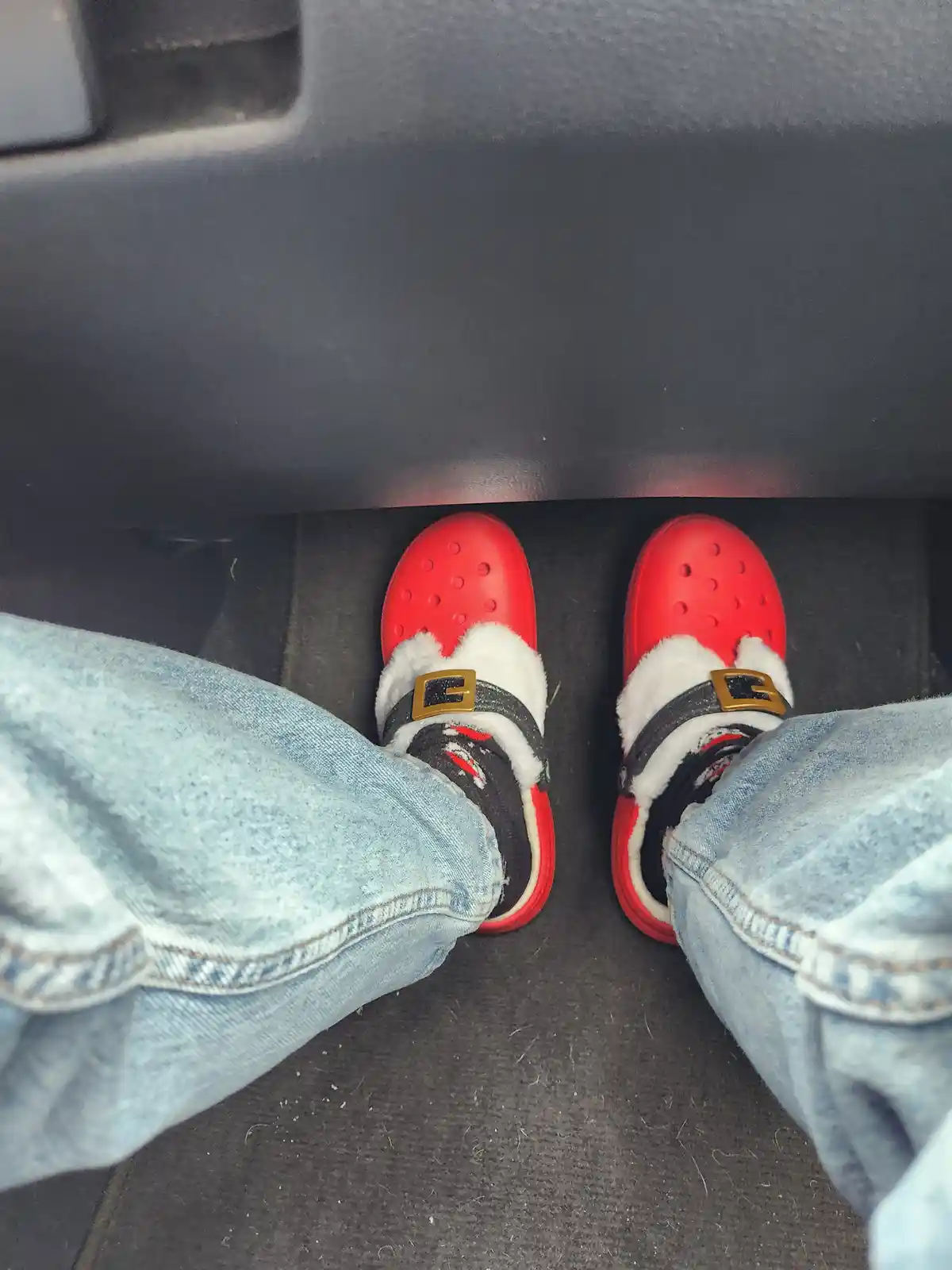 A first-person view from inside a car shows a person's feet wearing bright red, fuzzy, Santa-themed Crocs with white trim…