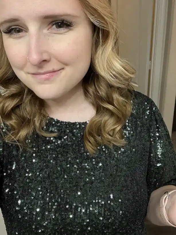 A close-up selfie of a woman with blonde wavy hair, wearing a black sequined top and a pink watch, smiling subtly while…