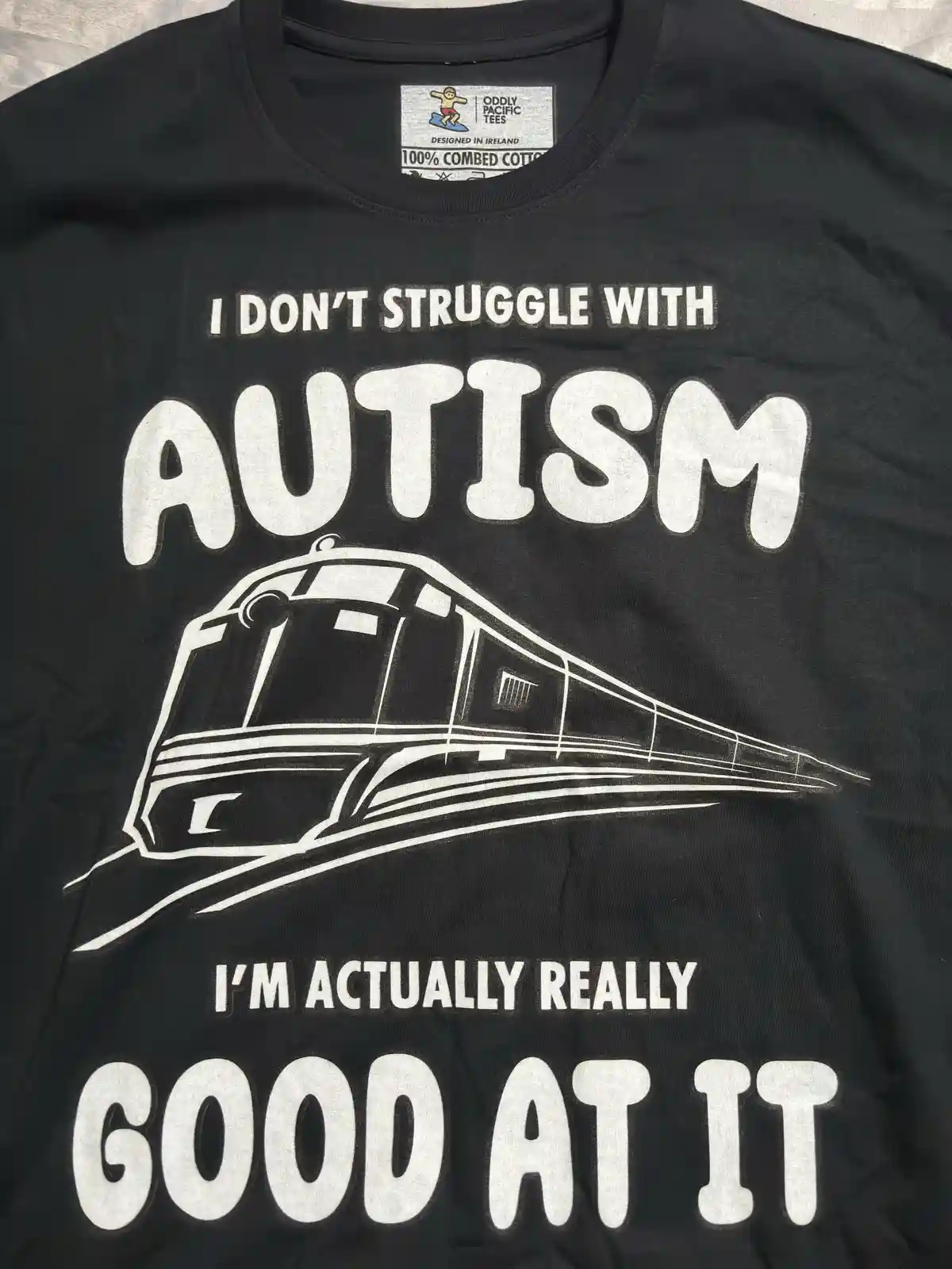 A black t-shirt displays white text stating 'I DON'T STRUGGLE WITH AUTISM I'M ACTUALLY REALLY GOOD AT IT', with a white…