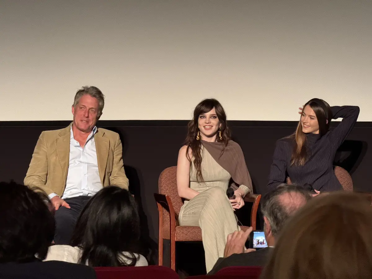 three people sit on a stage in front of an audience during a panel discussion. The person on the left, a man in a tan…