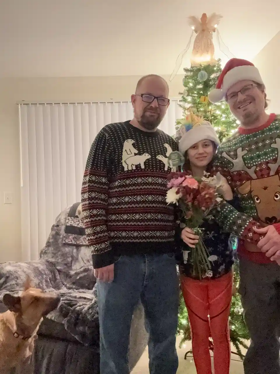 Three people pose for a family photo in front of a lit christmas tree, all wearing festive sweaters. The person on the left…