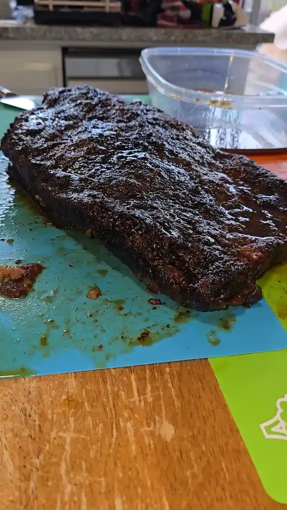 A large, darkly charred piece of meat, likely ribs or brisket, sits on a blue cutting mat with sauce drips, next to a clear…