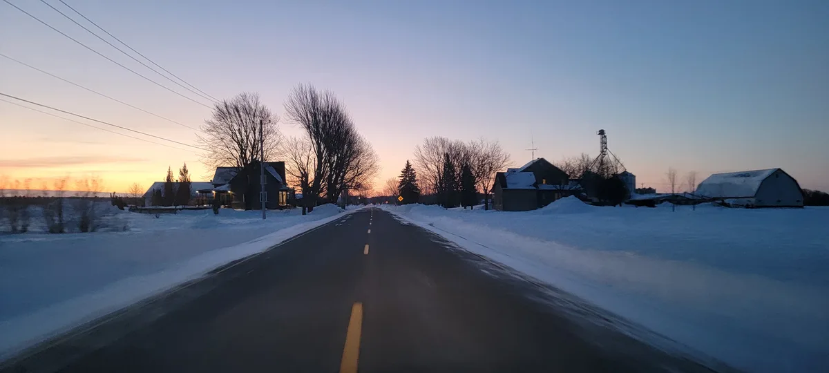 A point-of-view shot from a vehicle driving down a snow-covered rural road at dawn or dusk, with houses and barns visible on…