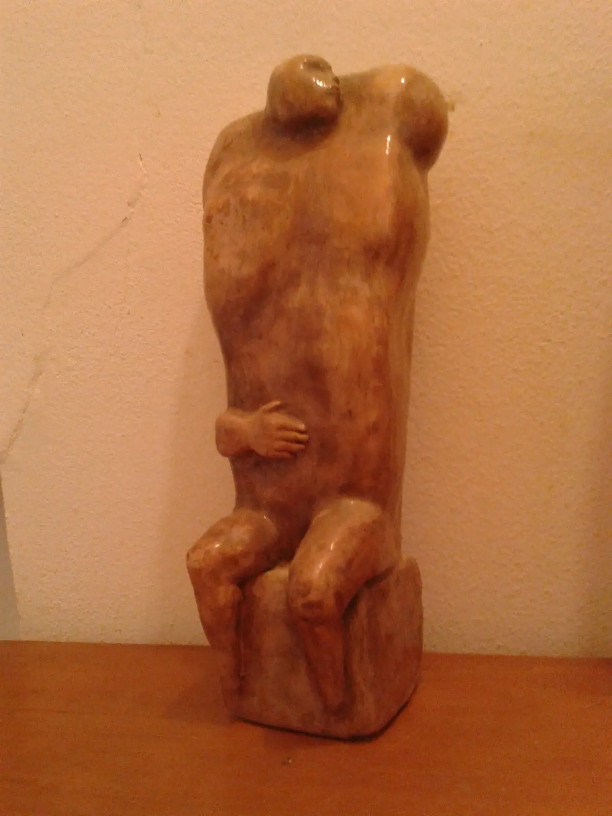 A brownish stone sculpture depicts two figures fused together, sitting on a pedestal, with one hand resting on the other's…