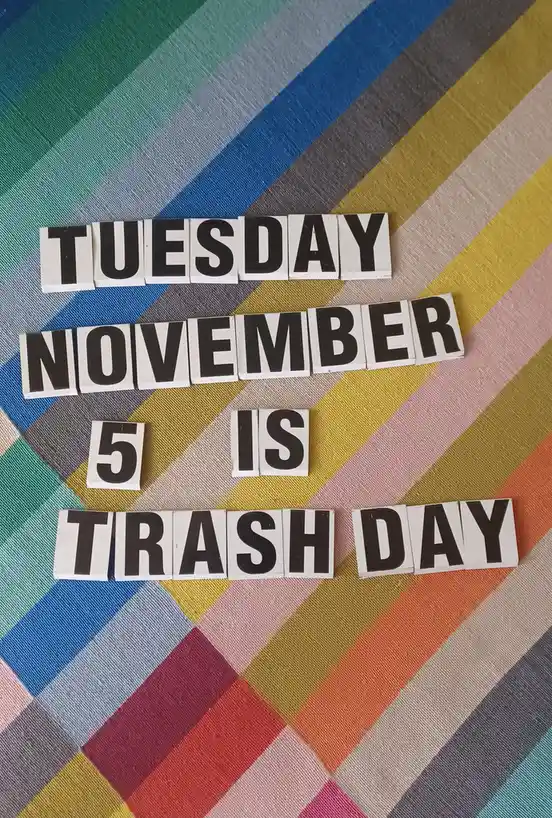 The image displays black-and-white letter tiles arranged on a colorful striped fabric background, spelling out 'TUESDAY…