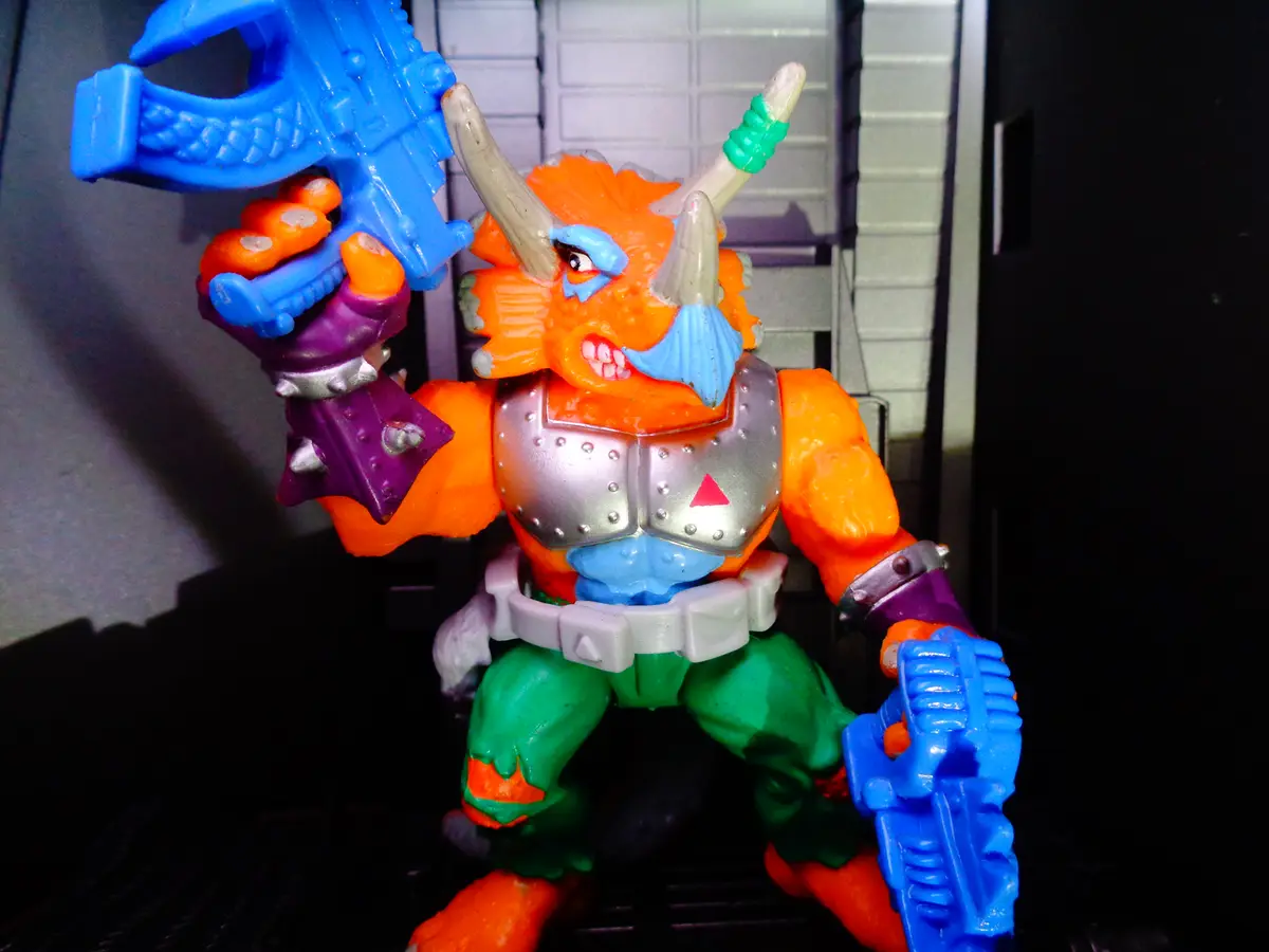 A brightly colored plastic action figure with orange skin, horns, and a blue mustache, wearing silver armor and holding two…