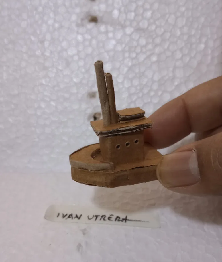 A hand holds a small, crudely crafted cardboard model of a ship with two smokestacks and three portholes. Below it, a white…