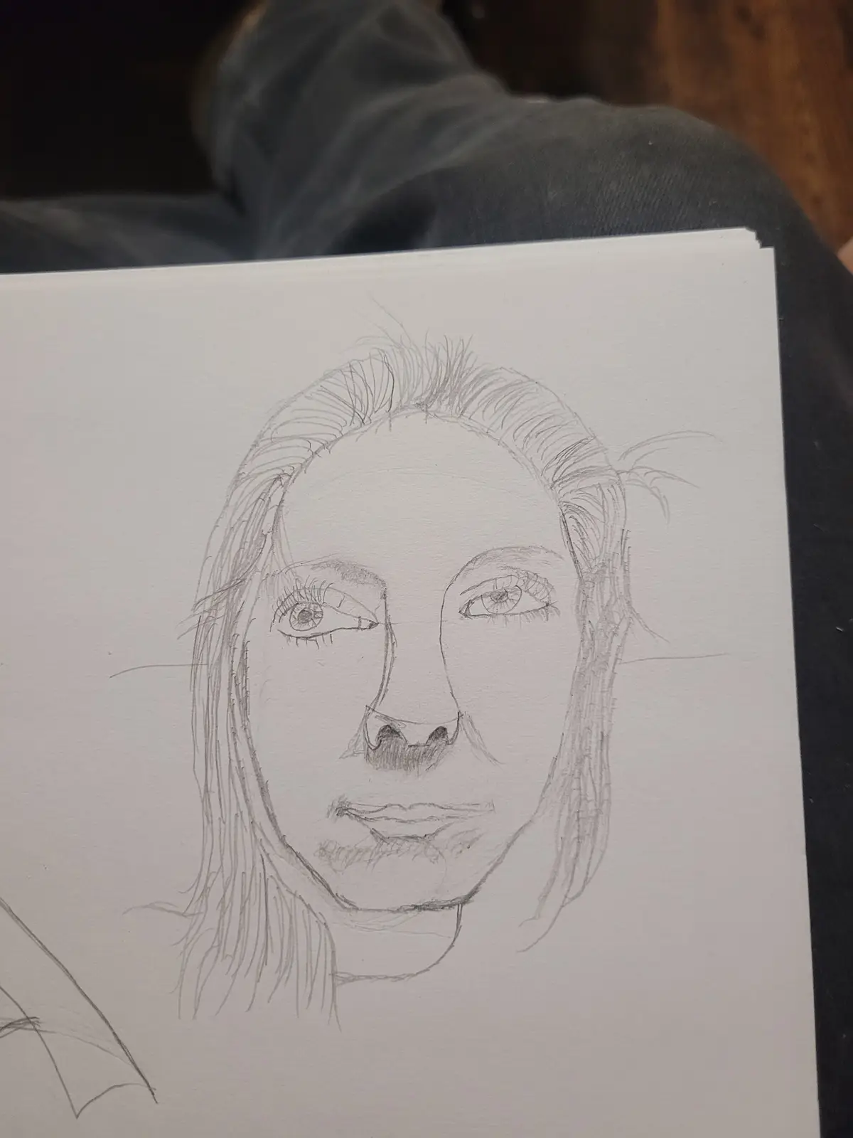 A pencil sketch on white paper shows a detailed portrait of a person with long hair, expressive eyes, and a neutral…