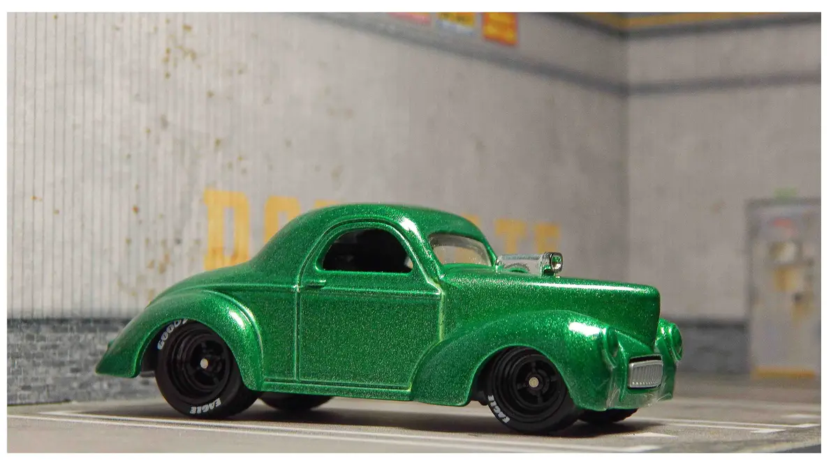 A shiny green die-cast model of a classic hot rod car is shown in profile, parked on a road with a blurred background. The…