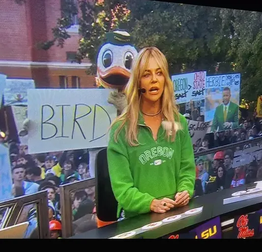 A woman in a green Oregon sweatshirt sits at a sports broadcast desk, looking surprised. Behind her, a large screen shows a…