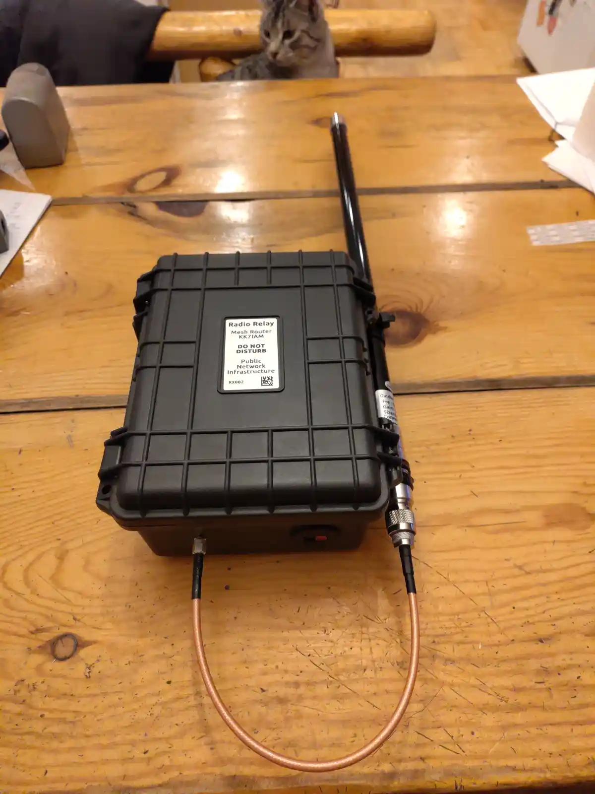 A black, rugged radio relay mesh router with a long antenna sits on a wooden table, featuring a label that reads 'Radio…