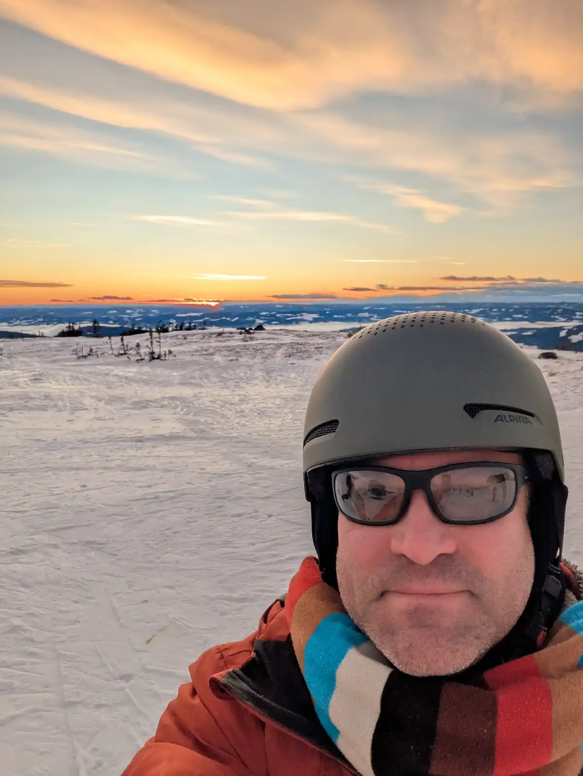 A man wearing a grey Alpina ski helmet, sunglasses, and a colorful plaid scarf stands in a snowy landscape at sunset. The…