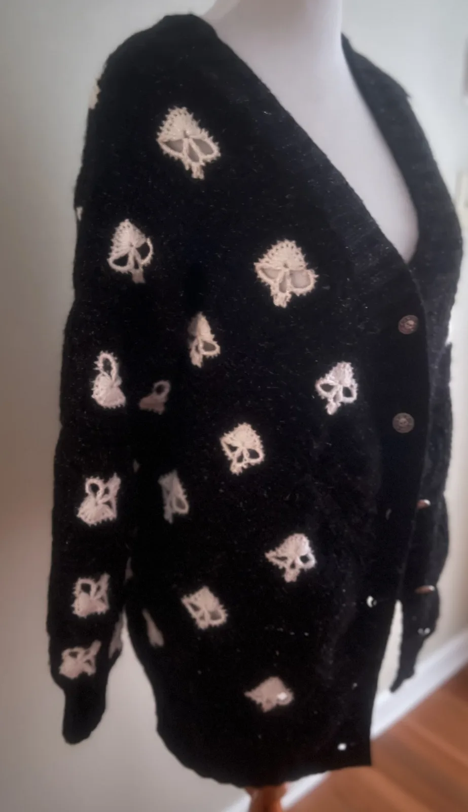 A black, fuzzy cardigan sweater with cream-colored skull patterns is displayed on a mannequin. The buttons are also…