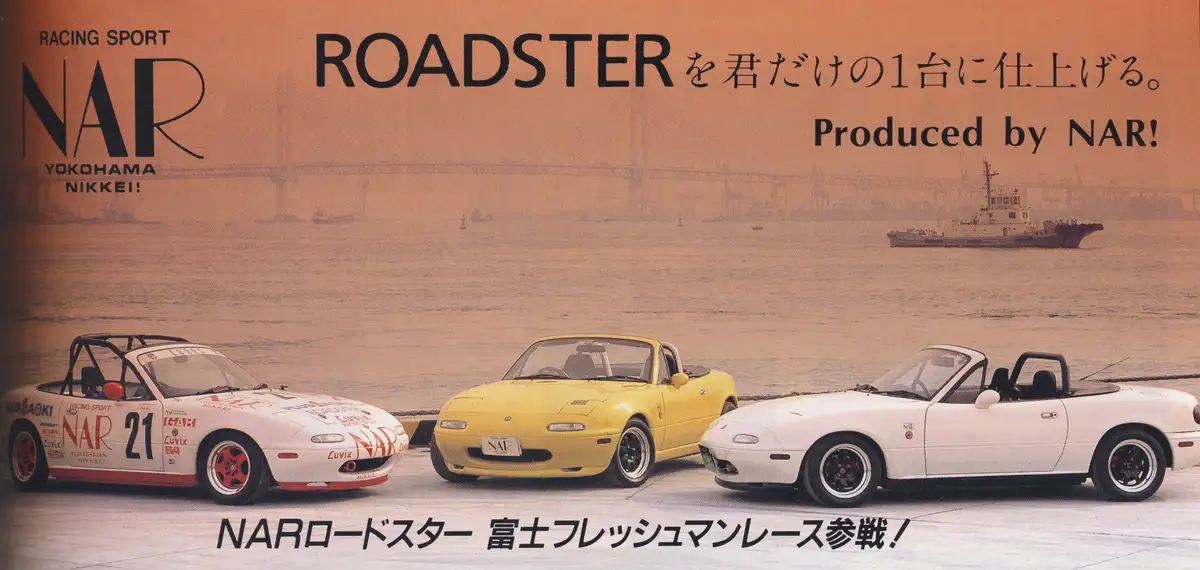 Three Mazda MX-5 Miata roadsters — one race-prepped white car with number 21, one yellow, and one white — are parked on a…