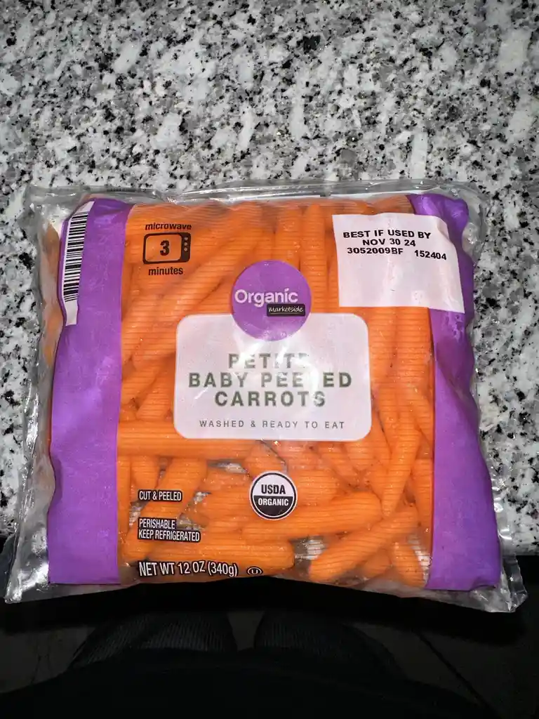 A close-up photo of a plastic bag of Marketside organic petite baby peeled carrots on a speckled countertop. The label shows…