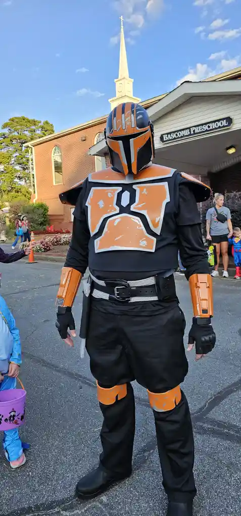 A person in a detailed Mandalorian armor costume with orange and black markings stands outside a building labeled 'BASCOMB…