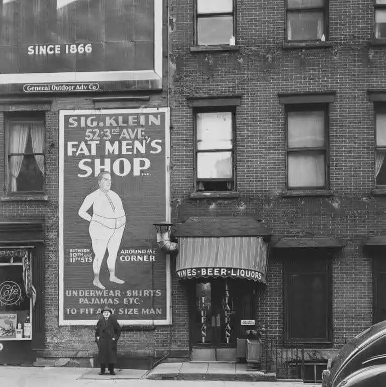 A black-and-white historical photo shows a large vintage billboard for 'Sig. Klein Fat Men's Shop' on a brick building…