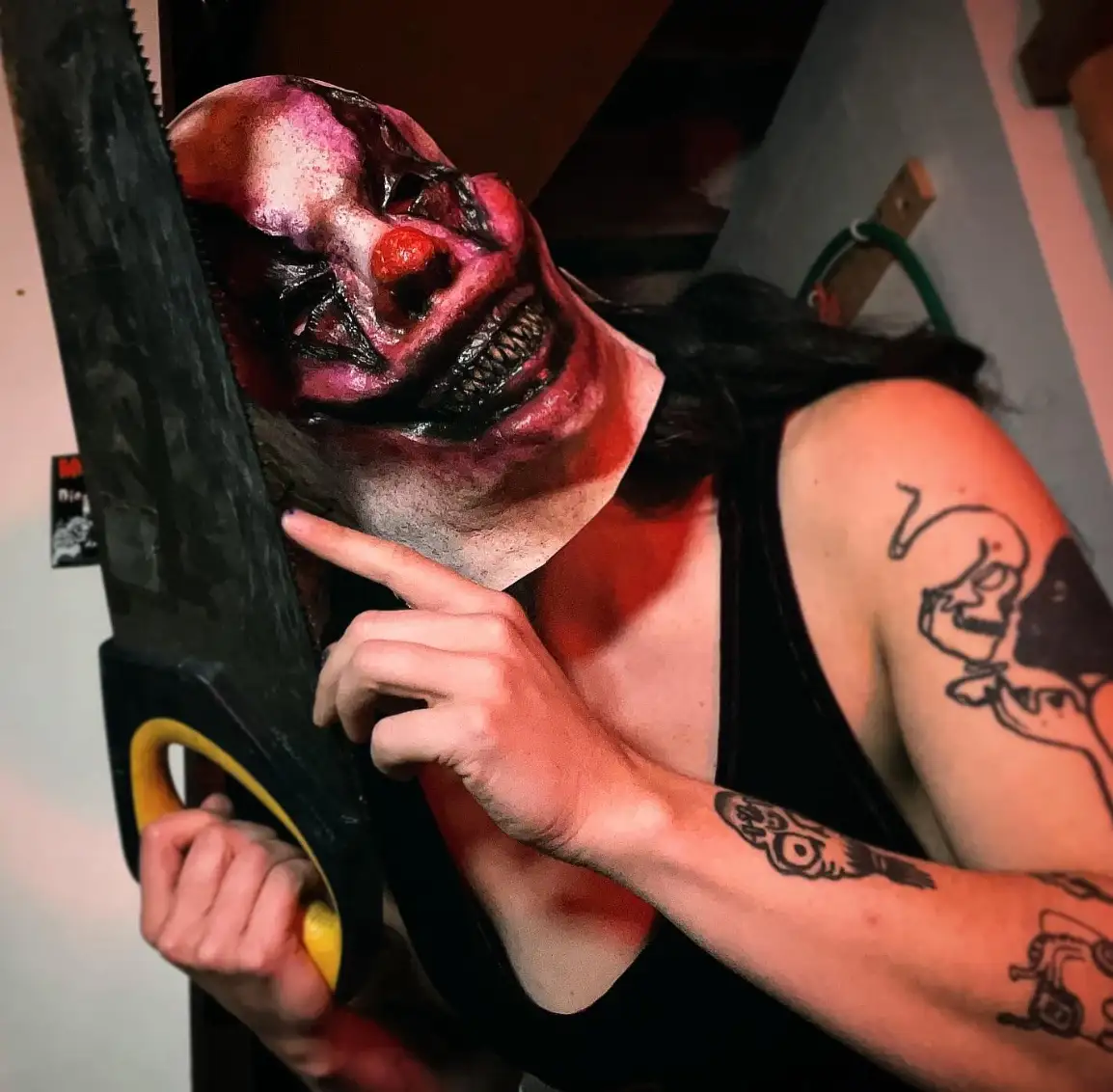 A person wearing a grotesque, bloody clown mask with a red nose and sharp teeth, holding a large handsaw. They point at the…