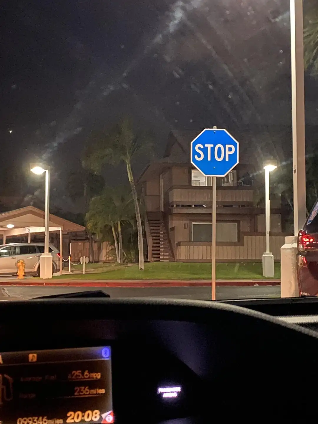 A nighttime photo taken from inside a car shows a standard blue octagonal stop sign in front of a residential building. The…