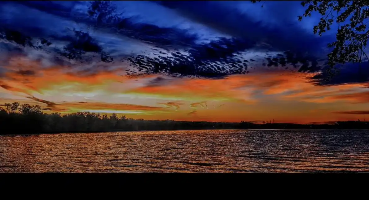 A dramatic sunset over a body of water with fiery orange and yellow clouds against a deep blue sky, silhouetted trees along…