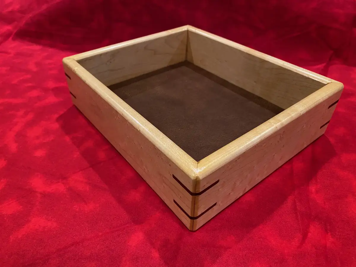 A light-colored wooden box with a dark brown felt interior sits on a red fabric surface. The box has visible dovetail joints…