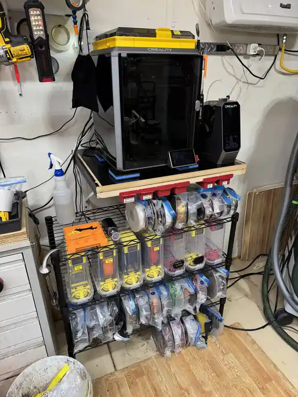 A well-organized 3d printing station features a Creality printer on a shelf above a black metal rack filled with neatly…