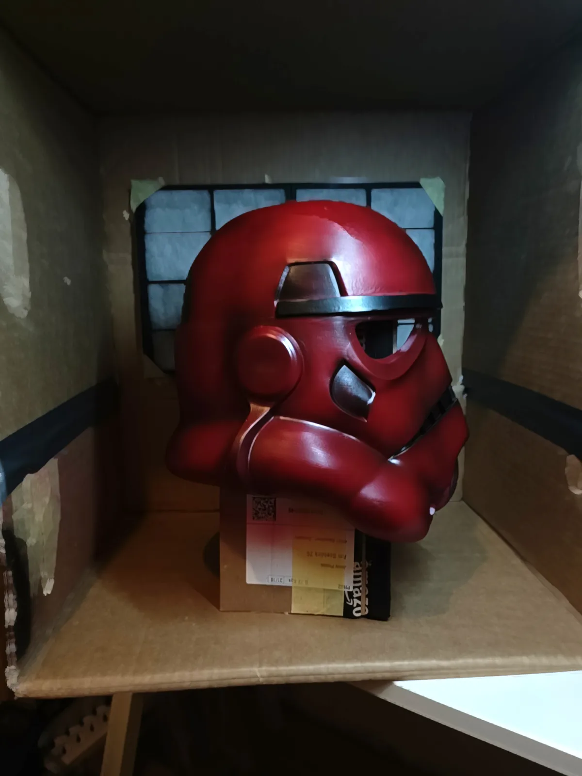 A glossy red stormtrooper helmet, resembling a Star Wars character, sits inside a cardboard box on top of an Amazon shipping…