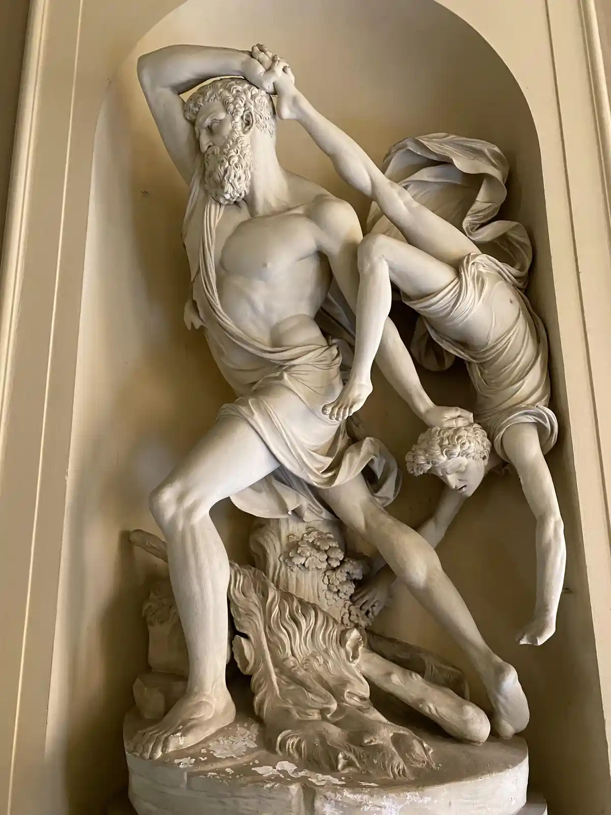 A classical marble sculpture depicts a bearded man in agony, clutching his head as a second figure pulls his hair, while a…