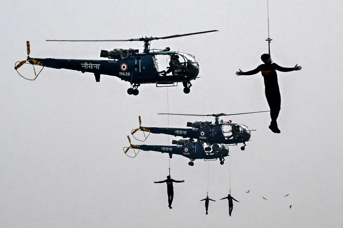 Three Indian Air Force helicopters fly in formation against a gray sky, each suspended by cables from the helicopters are…