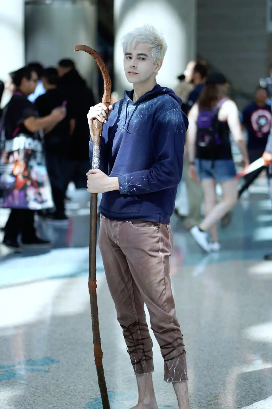 A young man with bleached blonde hair stands barefoot in a public space, holding a rustic wooden staff. He wears a dark blue…