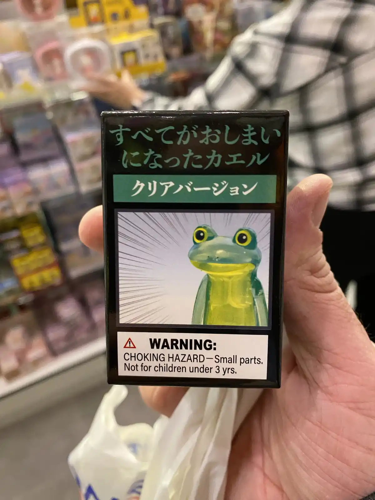 A hand holds a small toy box featuring a cartoon frog with a surprised expression, set against a white background with…
