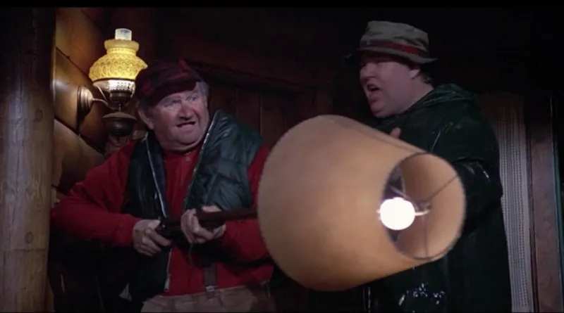 two men in a dimly lit wooden cabin, one holding a shotgun and the other holding a large lampshade with a lightbulb inside…