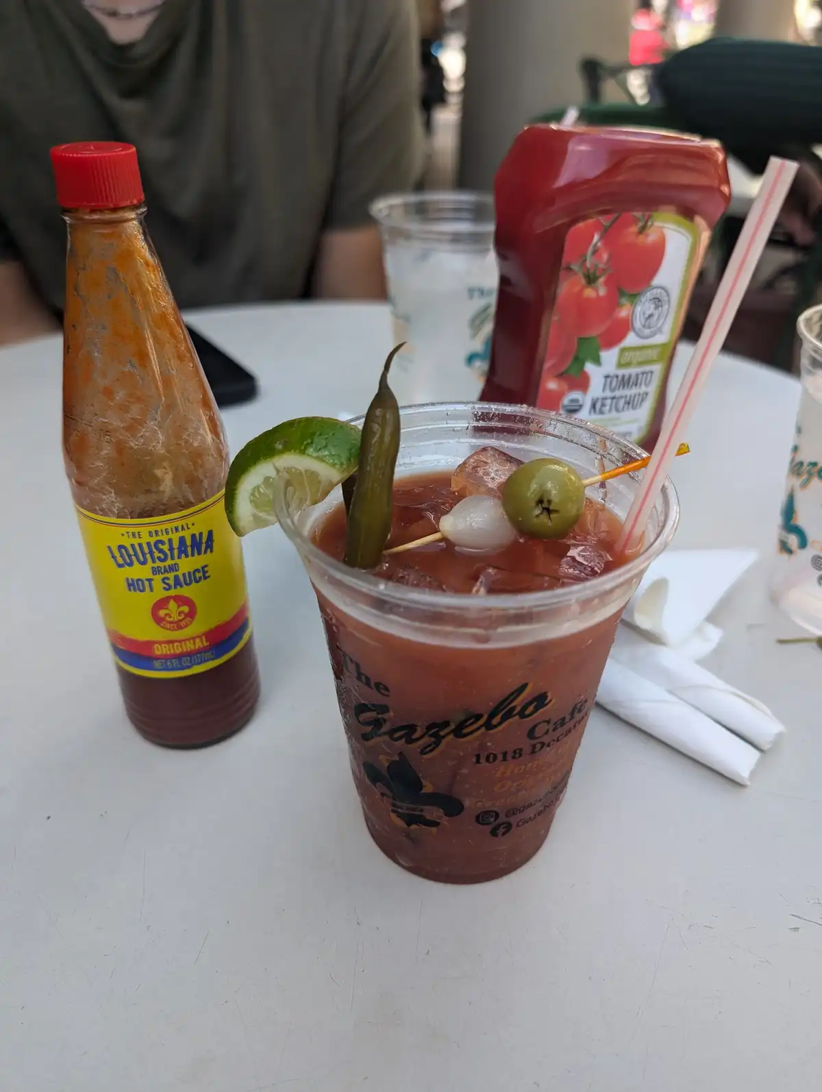a close-up photo shows a plastic cup of a red cocktail, likely a blood mary, garnished with a lime wedge, a pickle, an olive…
