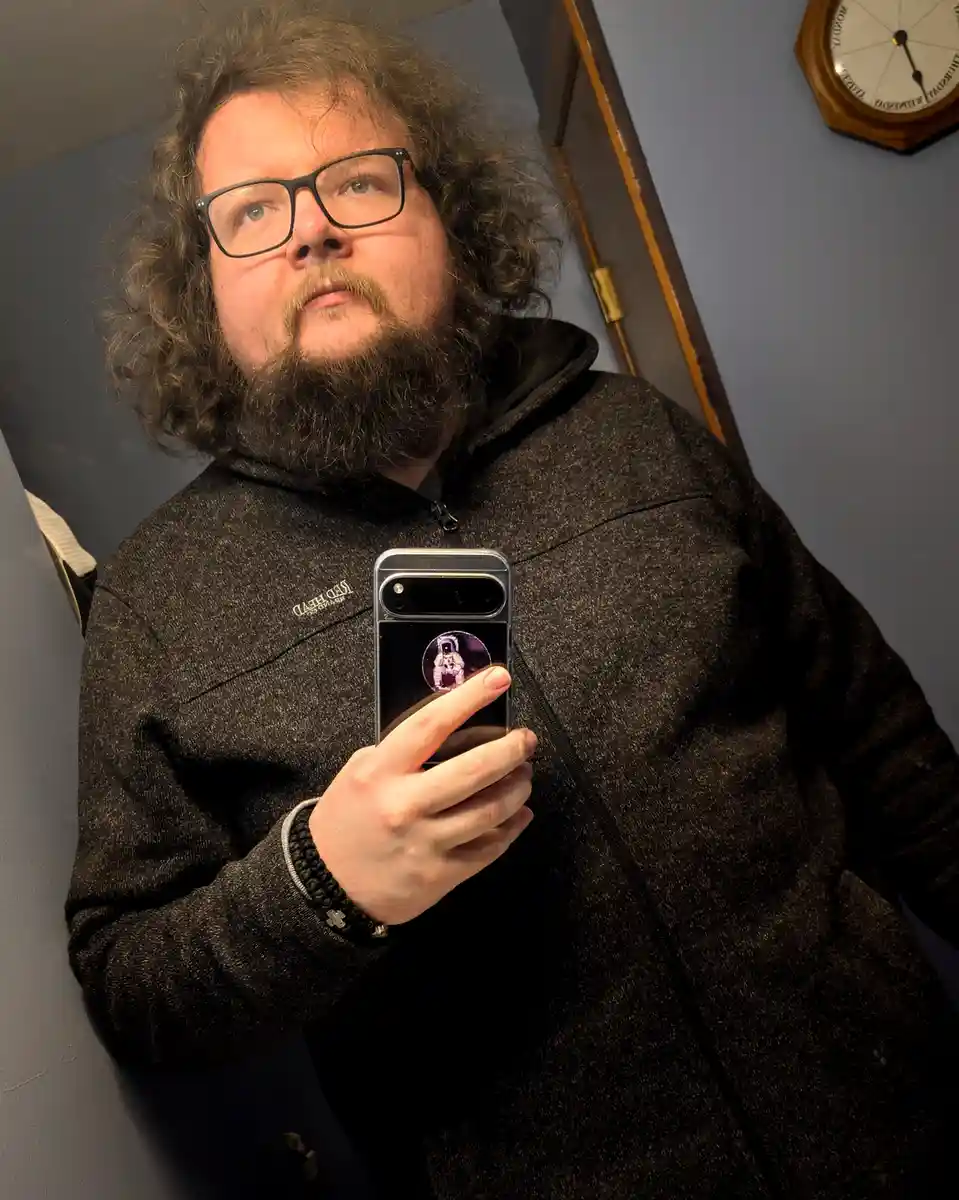 A man with curly hair, glasses, and a beard takes a mirror selfie holding a smartphone. He looks upward with a skeptical…