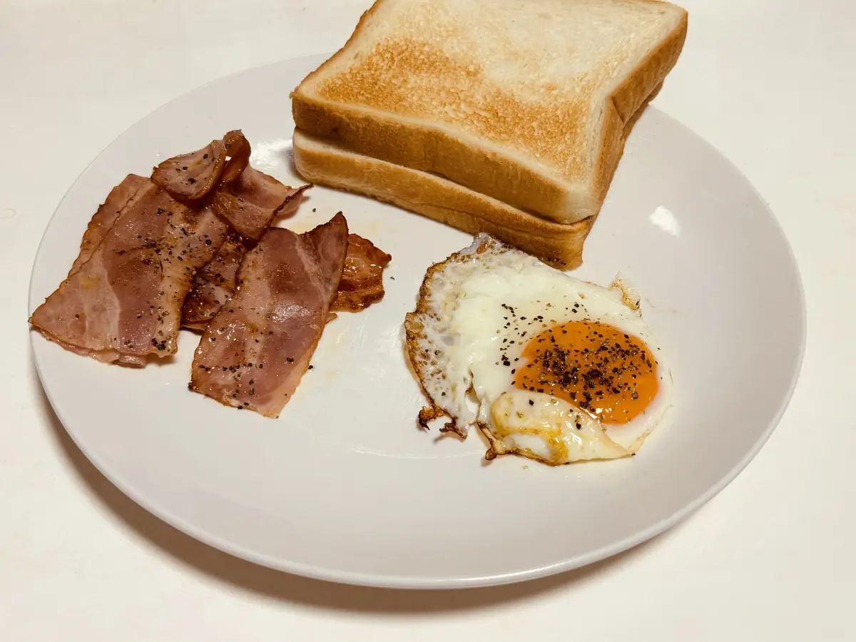 A plate of breakfast food including a fried egg, crispy bacon, and two slices of toast, all seasoned with black pepper. The…