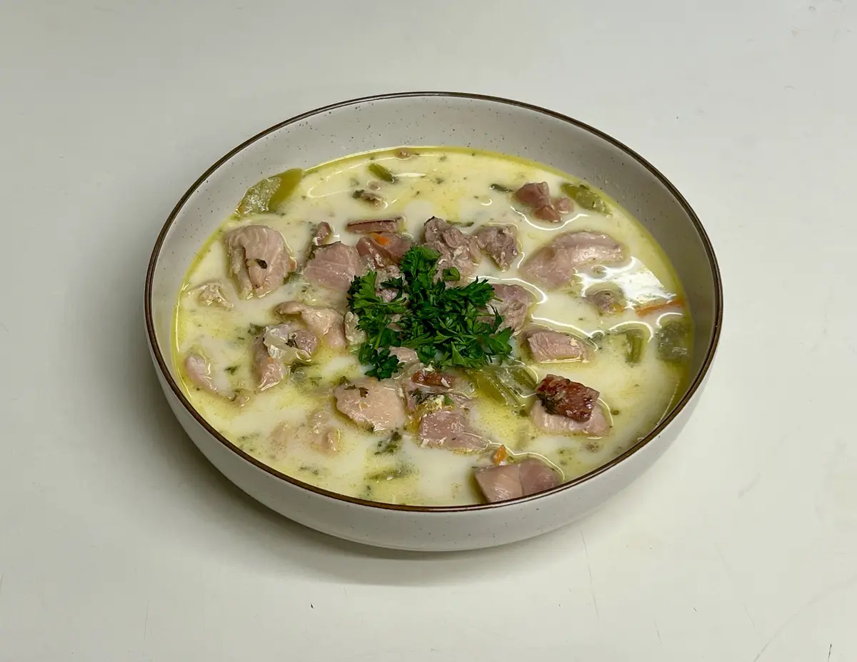 A bowl of creamy soup with chunks of ham, green vegetables, and a parsley garnish, presented in a speckled ceramic bowl on a…