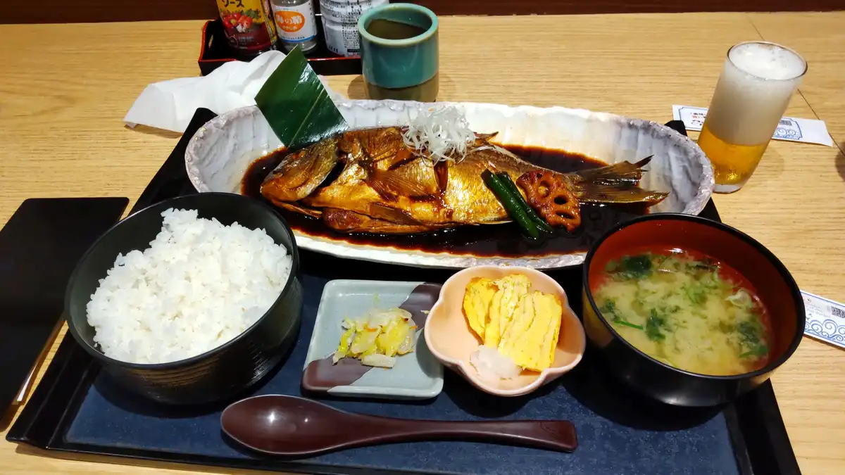 a full japanese meal is presented on a tray, featuring a whole fried fish in sauce, a bowl of white rice, miso soup, pickled…