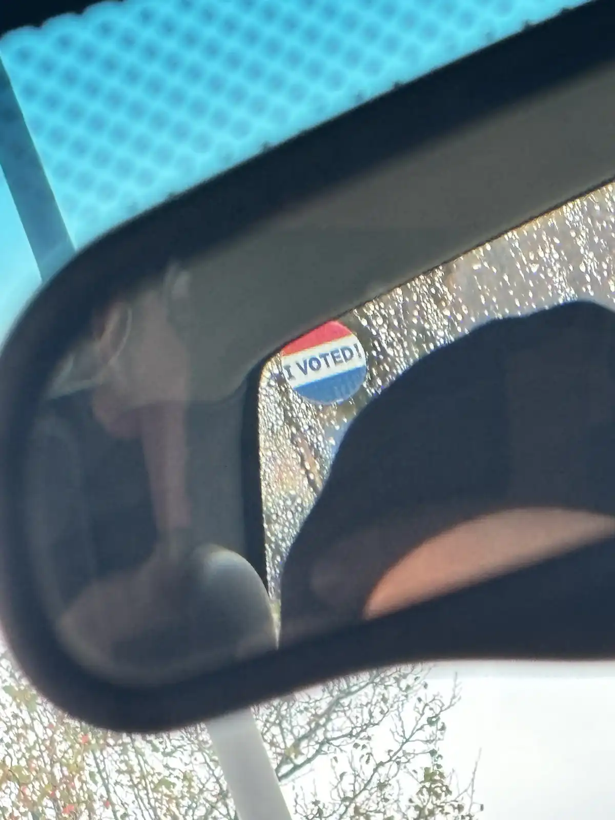 A close-up photo shows an 'I VOTED!' sticker stuck to the inside of a car's rearview mirror, with raindrops visible on the…