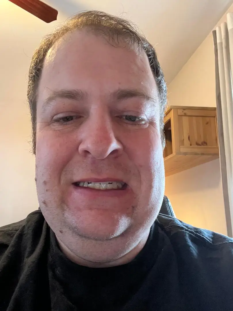 A close-up selfie of a man with a tired, slightly squinting expression, wearing a black shirt, with a wooden cabinet and…