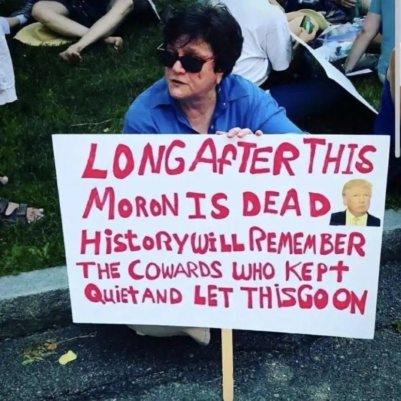 A person wearing sunglasses and a blue shirt sits on grass holding a handmade protest sign with red handwritten text. The…