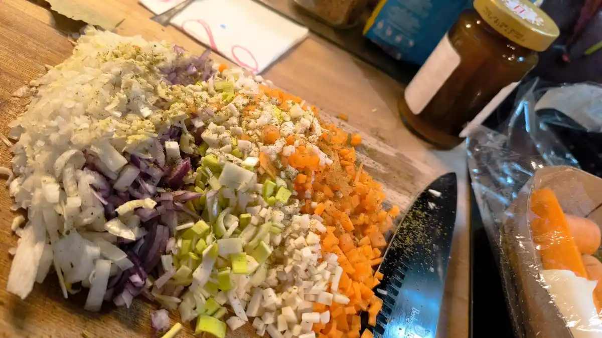 A wooden cutting board holds piles of finely chopped vegetables including white onions, red onions, leeks, and carrots, all…