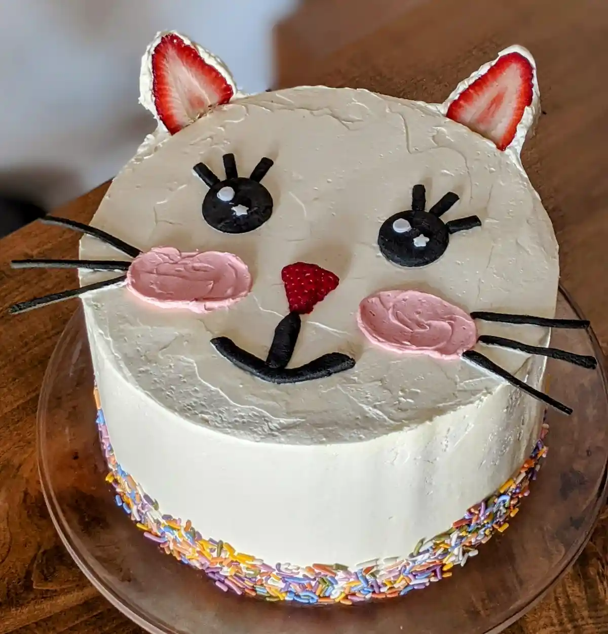 A round white-frosted cake decorated to resemble a cat's face, with strawberry slices forming ears, black icing for eyes and…