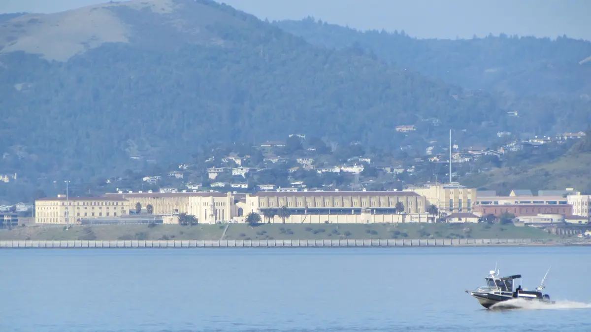 A wide shot shows a large, beige, institutional-style prison complex situated on a grassy shoreline next to a body of water…