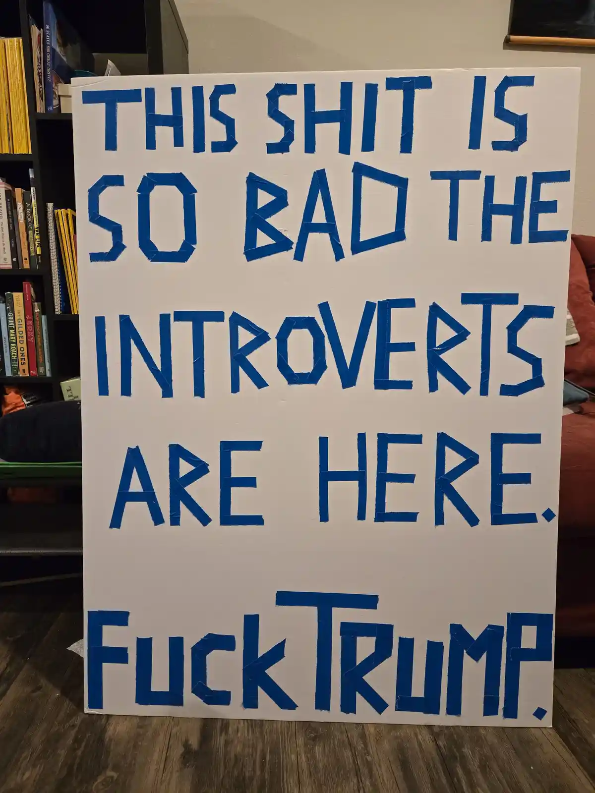 A handmade protest sign with blue tape letters on a white background stands indoors, reading 'THIS SHIT IS SO BAD THE…