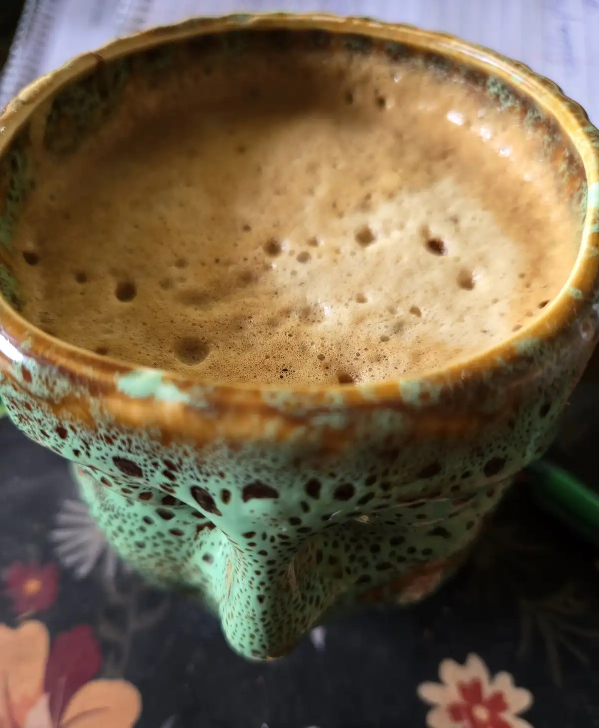 A close-up photo shows the top of a ceramic mug with a green and brown speckled glaze, filled with coffee that has a thick…