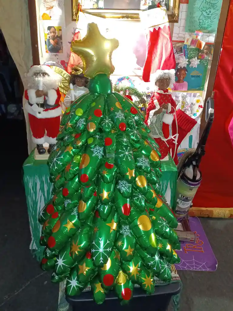 A large, green, inflatable Christmas tree made of balloons with red, gold, and white decorations sits on a table. It has a…