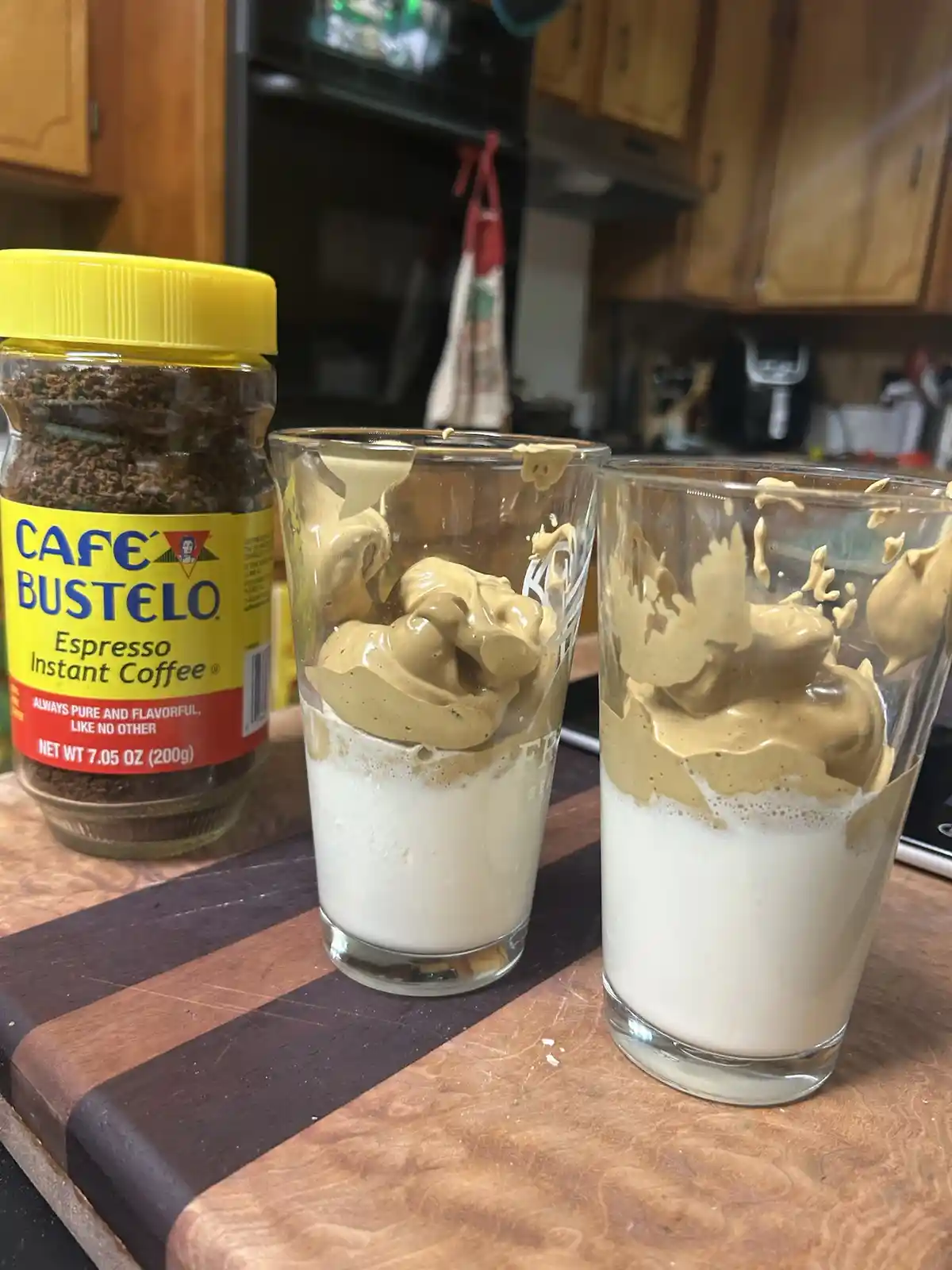 Two glasses sit on a wooden cutting board, each containing a white liquid base topped with a thick, light-brown, peanut…