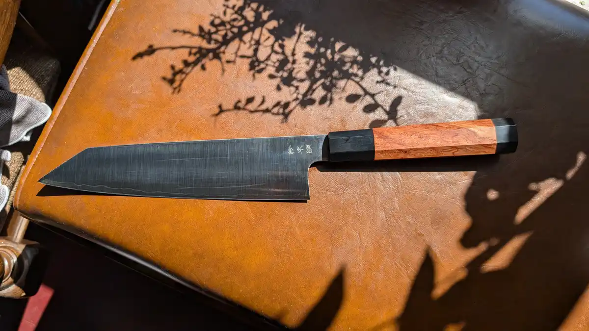 A high-quality Japanese chef's knife with a wooden handle and black bolster rests on a brown leather surface, illuminated by…